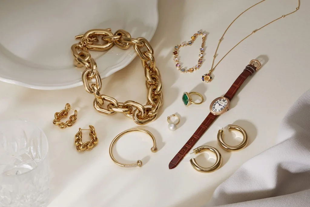 چندین دستبند، گوشواره و گردنبند زیبا روی سطح سفید به همراه یک ساعت کوچک زنانه با بند چرم قهوه‌ای