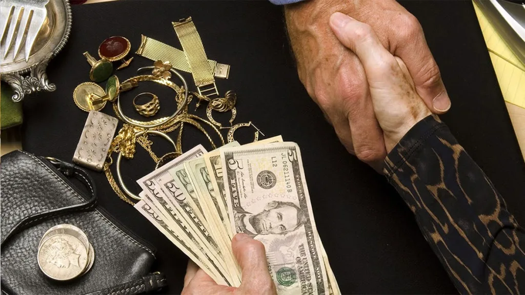 نمای بالای دست دادن فروشنده طلا و خریدار با تعدادی اسکناس دلاری در دست فروشنده و مقداری طلاهای زنانه که از یک کیف چرم بیرون آورده شده است.