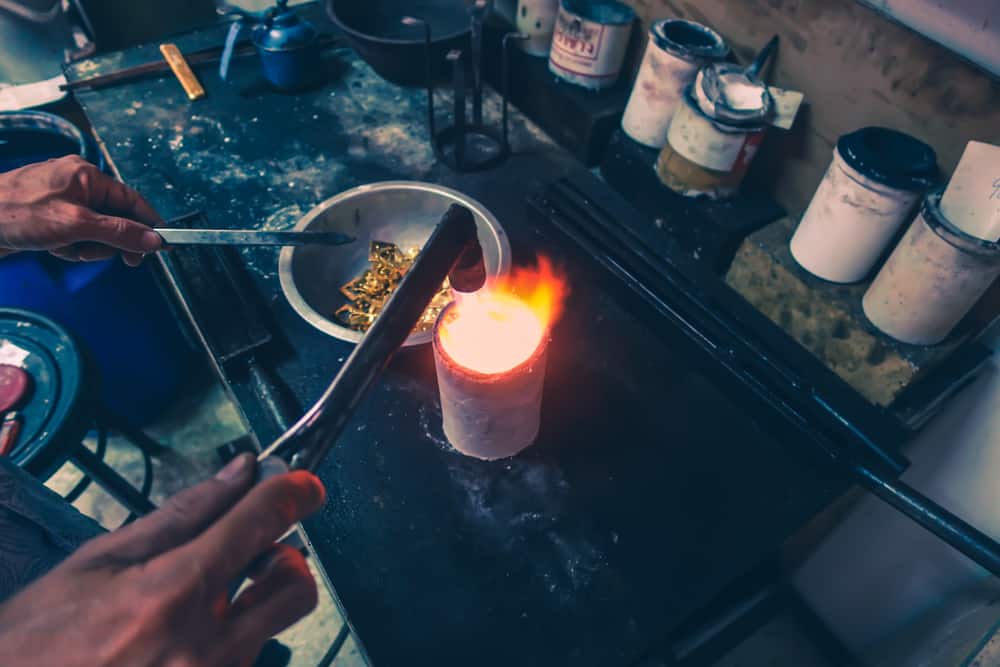  نمای بالاسری از دست یک طلاساز که با انبر دست طلا را از ظرف آهنی بر می‌دارد و روی کوره استوانه‌ای گرم می‌کند