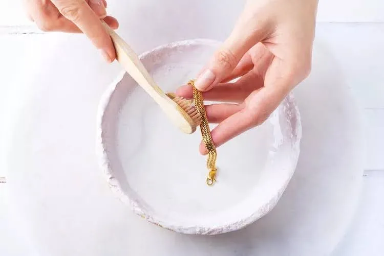 نمای بالا از یک کاسه سفید حاوی محلول آب و تاید و دستان زنی در حال کشیدن مسواک چوبی روی دستبند طلا
