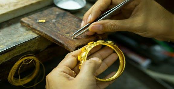 نمای بالا از دستان یک طلاساز در حال قرار دادن نگین روی یک دستبند طلای زنانه
