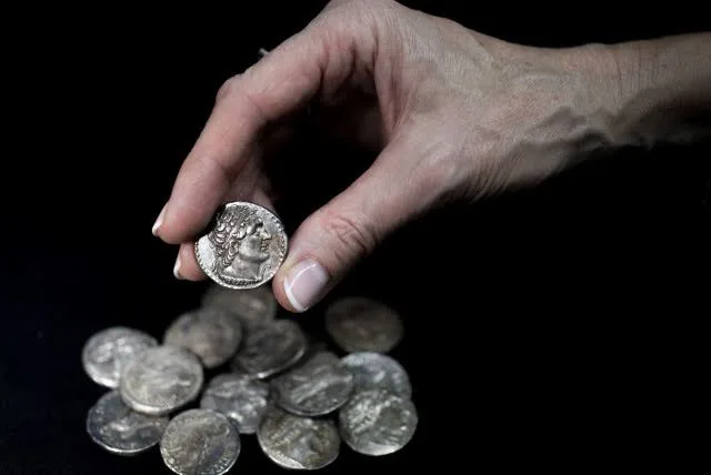 نمای روبه‌رو از تعدادی سکه نقره در پس زمینه‌ای سیاه و دست راست یک سرمایه‌گذار با یک سکه نقره خارجی میان انگشتان سبابه و شست