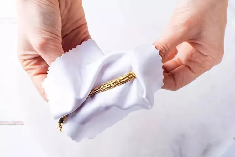 نمای روبه‌رو از دستان زنی در حال خشک کردن دستبند طلای خود با یک پارچه سفید نرم