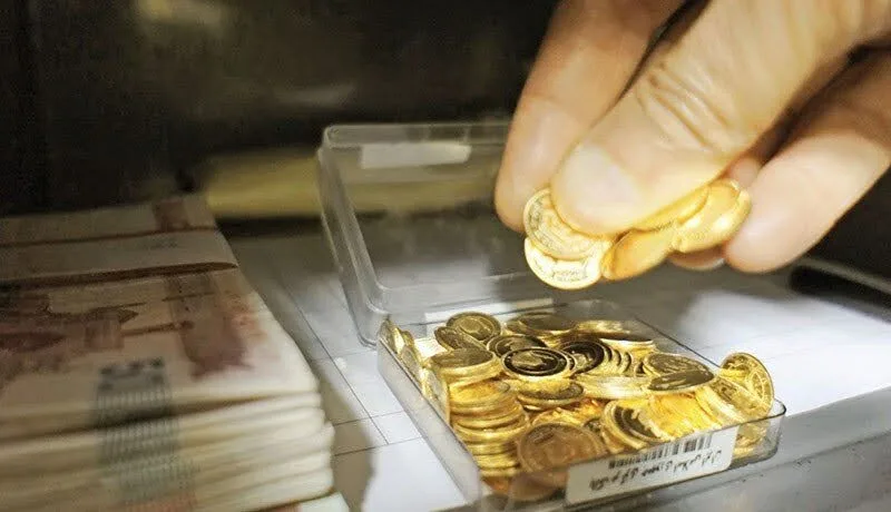 دست طلافروش مرد در حال برداشتن تعدادی سکه از یک جعبه پر از سکه گرمی در کنار تاول‌های ۵۰ هزار تومانی