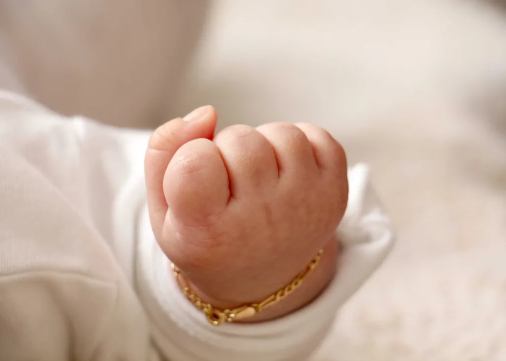 نمای روبرو از دست گره کرده نوزاد در حالت خوابیده با دستبند طلا و لباس سفید