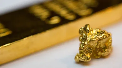 تصویر یک تکه طلا به شکل سنگ روی یک میز سفید و عکس یک شمش طلا در پس زمینه به صورت تار