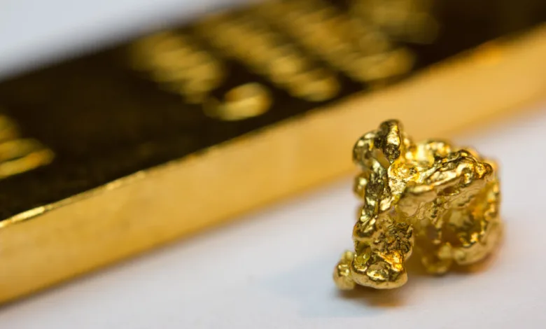 تصویر یک تکه طلا به شکل سنگ روی یک میز سفید و عکس یک شمش طلا در پس زمینه به صورت تار