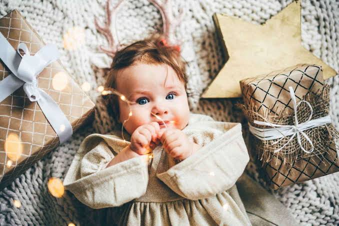 نمای بالا از نوزاد دختری با چشمان آبی روی یک زیرانداز بافتنی طوسی و جعبه‌های کادو در اطرافش