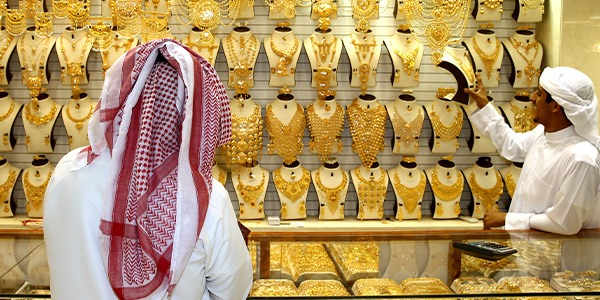 نمای روبه‌رو از پیشخوان یک مغازه طلافروشی در عربستان که فروشنده در حال معرفی و نشان دادن یک گردنبند طلا به خریدار عربستانی است.
