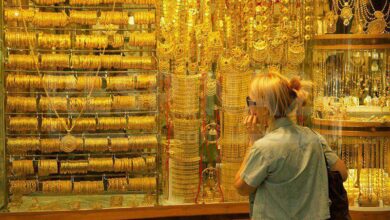 نمای روبه‌رو از پشت شیشه مغازه طلافروشی با انواع طلا و زن مسن در حال تماشای طلاها از پشت شیشه