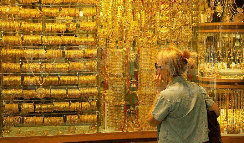 نمای روبه‌رو از پشت شیشه مغازه طلافروشی با انواع طلا و زن مسن در حال تماشای طلاها از پشت شیشه